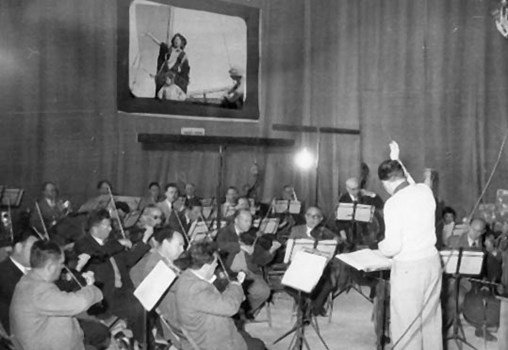 משה וילנסקי מנצח באולפן הקלטות על ביצוע המוזיקה לסרט "באין מולדת" (מוקרן ברקע), 1958​ (ארכיון משה וילנסקי, MUS 0069)